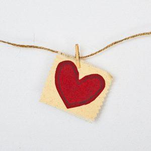 A photo of a piece of twine going across the middle of a white background and attached to the twine, with a tiny clothespin, is a piece of beige paper with a heart in the center to represent Valentines Day.