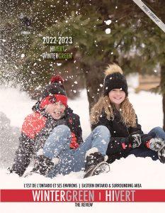 This is an image for the front cover of the 2022-23 WinterGreen Magazine one of the Prescott-Russell Tourism Magazine Publications The Review does each year. It has two children a boy and a girl in winter coats and hats sitting in the snow having fun and smiling